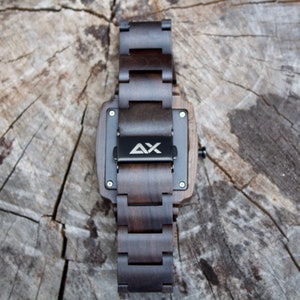 K&ouml;nnte beinhalten: Eine dunkelbraune Holzuhr mit schwarzem Zifferblatt und einem Metallband. Die Uhr hat ein quadratisches Zifferblatt und auf der R&uuml;ckseite der Uhr sind die Buchstaben "AX" eingraviert.