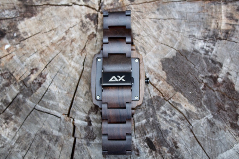 K&ouml;nnte beinhalten: Eine dunkelbraune Holzuhr mit schwarzem Zifferblatt und einem Metallband. Die Uhr hat ein quadratisches Zifferblatt und auf der R&uuml;ckseite der Uhr sind die Buchstaben "AX" eingraviert.