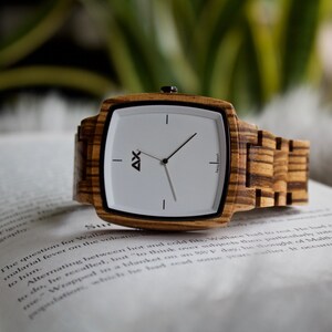 K&ouml;nnte beinhalten: Eine Armbanduhr aus Holz mit wei&szlig;em Zifferblatt und schwarzen Zeigern. Die Uhr hat ein quadratisches Zifferblatt und ein braunes Holzband.