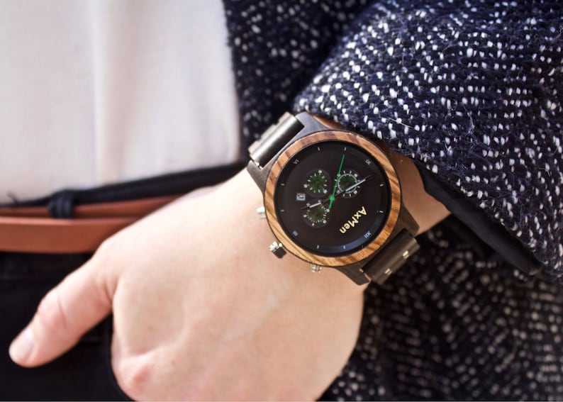 Puede incluir: Un reloj de pulsera de madera negro y marr&oacute;n con una esfera de cron&oacute;grafo. La esfera del reloj tiene un fondo negro con n&uacute;meros blancos y detalles verdes. El nombre de la marca "AXMEN" est&aacute; impreso en la esfera del reloj.