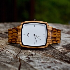 K&ouml;nnte beinhalten: Eine quadratische Holzuhr mit wei&szlig;em Zifferblatt und schwarzen Zeigern. Die Uhr hat ein Holzband mit einem gestreiften Muster.