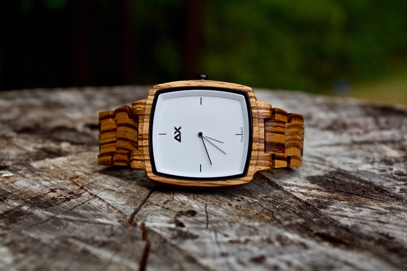 K&ouml;nnte beinhalten: Eine quadratische Holzuhr mit wei&szlig;em Zifferblatt und schwarzen Zeigern. Die Uhr hat ein Holzband mit einem gestreiften Muster.