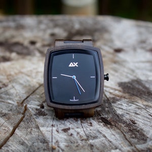 K&ouml;nnte beinhalten: Eine quadratische Holzuhr mit schwarzem Zifferblatt und wei&szlig;en Zeigern. Die Uhr hat ein schwarzes Lederarmband und der Text "AX Swiss Made" ist auf dem Zifferblatt aufgedruckt.