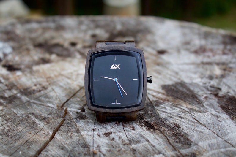 K&ouml;nnte beinhalten: Eine quadratische Holzuhr mit schwarzem Zifferblatt und wei&szlig;en Zeigern. Die Uhr hat ein schwarzes Lederarmband und der Text "AX Swiss Made" ist auf dem Zifferblatt aufgedruckt.