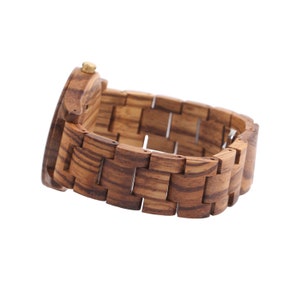 Puede incluir: Un reloj de pulsera de madera con una banda de madera marr&oacute;n y una corona de color dorado. La esfera del reloj no es visible en la imagen.
