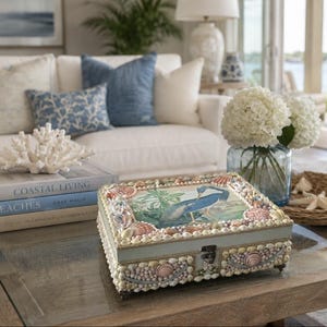 May include: A rectangular light blue wooden box decorated with various small seashells along the edges and lid. The top of the box features a central illustration of a blue heron standing among green foliage, secured with a metal latch.