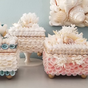 May include: Three decorative boxes made of seashells. The boxes are square and have a layered design. The top box is white with a coral design, the middle box is white with a blue trim, and the bottom box is pink with a white trim.