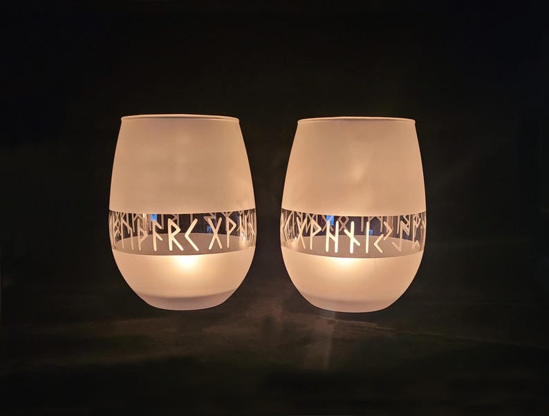 K&ouml;nnte beinhalten: Zwei Kerzenhalter aus mattiertem Glas mit einem silbernen Band, das mit Runen verziert ist. Die Kerzen sind angez&uuml;ndet und leuchten.