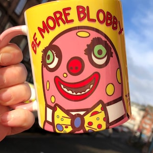 May include: A yellow ceramic mug with the words "BE MORE BLOBBY" in red. The mug features a pink clown face with green eyes, a red nose, and a yellow bow tie. The handle is pink. A fun, quirky design.