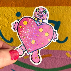 Puede incluir: Una pegatina rosa con un personaje en forma de corazón con lunares amarillos y una pajarita. El personaje tiene una cara sonriente y los brazos extendidos. La pegatina tiene un acabado brillante.