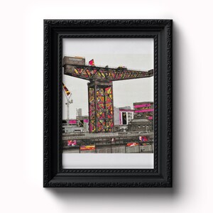 May include: A black framed print of a colourful, geometrically patterned crane structure with the text "Clydeport" on the side. The crane is set against a grey sky and a cityscape.