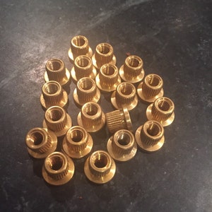 May include: A group of 20 gold-colored metal nuts with a threaded center hole. The nuts are arranged in a loose pile on a dark gray surface.