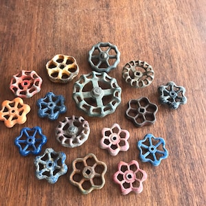 May include: A collection of 15 vintage metal water valve handles in various colors and designs. The handles are arranged in a circular pattern on a wooden surface.