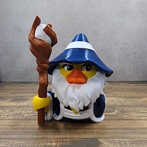 May include: A blue and white duck wearing a wizard hat and holding a brown staff with a white ball on top. The duck has a white beard and a yellow beak.