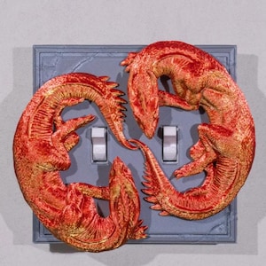 May include: A light switch cover with two red and gold dragons sculpted around it. The dragons are curled up around the switch, with their tails intertwined. The switch cover is made of a gray material.