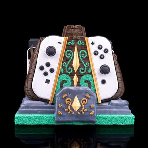 May include: A green and brown Nintendo Switch charging stand with a decorative design featuring a diamond and swirls. The stand is designed to hold two controllers and the Switch console.