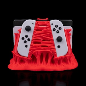 May include: A red 3D printed stand with a textured surface holds two white Nintendo Switch controllers. The stand is designed to resemble a flame.