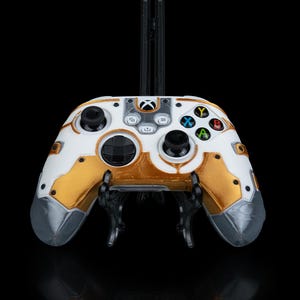 May include: A white and gold Xbox controller with a custom design. The controller is on a black stand.