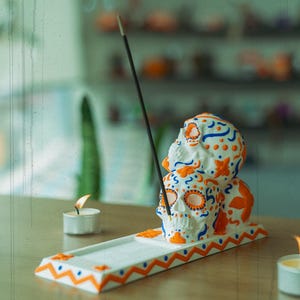 May include: White ceramic incense burner shaped like three stacked sugar skulls with orange and blue floral designs. The burner has a white and orange zig-zag pattern on the base.
