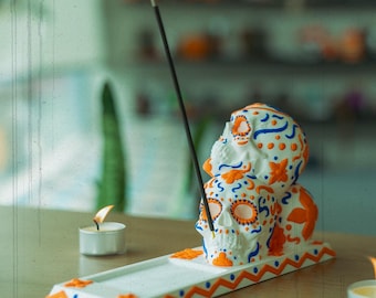 May include: White ceramic incense burner shaped like three stacked sugar skulls with orange and blue floral designs. The burner has a white and orange zig-zag pattern on the base.