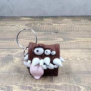 May include: A brown and white 3D printed keychain shaped like a treasure chest with a toothy grin and a pink tongue sticking out. The keychain has a silver ring attached to the top.