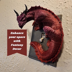Puede incluir: Una figurita de dragón rojo está unida a un interruptor de luz. El dragón está enroscado con su cola envuelta alrededor del interruptor. El texto "Enhance your space with Fantasy Decor" es visible en la imagen.
