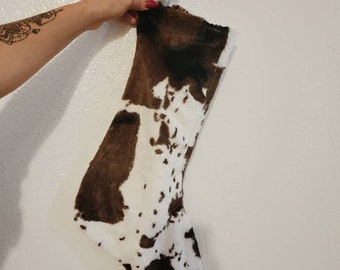 Könnte beinhalten: Ein Weihnachtsstrumpf mit Kuhfellmuster. Der Strumpf ist überwiegend weiß mit braunen Flecken und Flecken und hat eine Schlaufe zum Aufhängen. Das Kunstfellmaterial verleiht ihm ein weiches, strukturiertes Aussehen. Für die Feiertage entworfen.