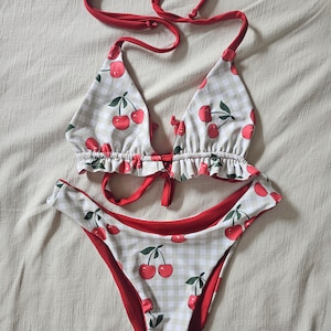 May include: A two-piece bikini set with a cherry print. The top is a triangle style with red ties, and the bottoms are high-cut. The fabric features a white and tan gingham pattern with red cherries and green leaves.