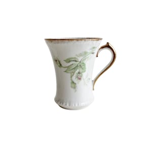 May include: White porcelain teacup with a gold handle and rim. The cup features a delicate floral design in green and pink. The handle is curved and elegant, and the rim has a gold trim.