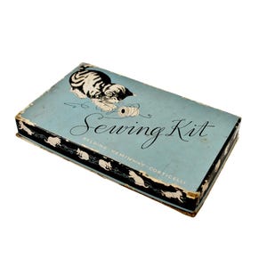 Puede incluir: Una caja de costura rectangular de color azul claro con una ilustración en blanco y negro de un gatito jugando con un carrete de hilo. El texto Sewing Kit está impreso en letra cursiva negra, con Belding-Heminway-Corticelli debajo. Los lados son negros con siluetas de gatos blancos.