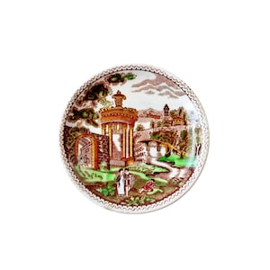 May include: A round decorative plate with a detailed landscape scene in brown, white, and green. The plate features a building with a domed roof, a bridge, and figures. The rim has a scalloped edge.