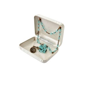 Puede incluir: Una caja de joyería rectangular plateada abierta, con un collar de cuentas azul claro y un pequeño colgante redondo en su interior. La caja está sobre un fondo blanco.