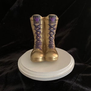Könnte beinhalten: Ein Paar goldfarbene Stiefel mit violetten Schnürsenkeln und einem violetten Logo an der Seite. Die Stiefel stehen auf einem weißen Sockel.