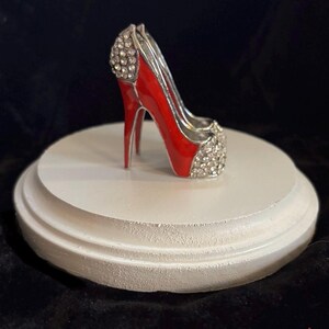 May include: A red high heel shoe with silver accents and rhinestones on a white pedestal.