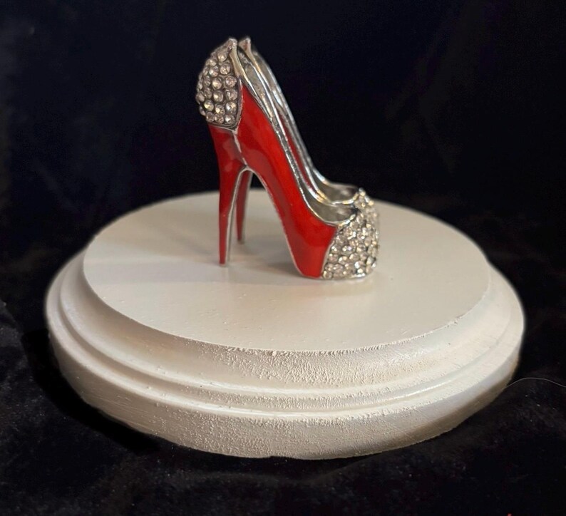 May include: A red high heel shoe with silver accents and rhinestones on a white pedestal.