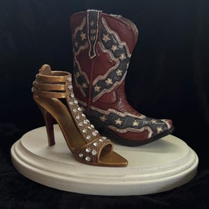 May include: A white cake topper featuring a brown cowboy boot with a Confederate flag design and a gold high-heeled shoe with rhinestones.