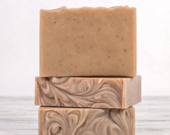May include: A stack of three rectangular bars of soap. The top bar is a solid light tan color with small embedded particles. The bottom two bars feature a swirling marbled pattern in shades of light brown, beige, and cream throughout the surface.