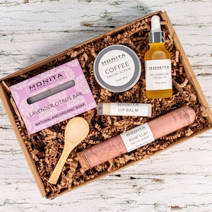 May include: A gift box containing Monita Natural Care products: a lavender citrus soap bar, a 37 g coffee facial scrub, a 30 ml rejuvenating facial oil, a lip balm, a rose clay facial mask, and a small wooden spoon nestled in brown paper.