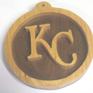 May include: A wooden ornament with the letters "KC" carved into it. The letters are made of light wood and are set against a dark wood background.