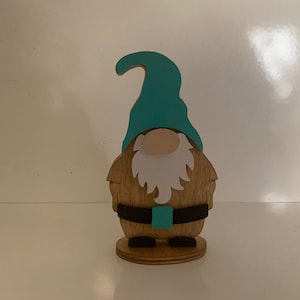 May include: A wooden gnome figurine with a teal hat, white beard, and black belt. The gnome is standing on a wooden base.