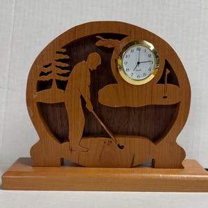 May include: A wooden clock with a golfer silhouette and a round clock face. The clock is on a wooden base.