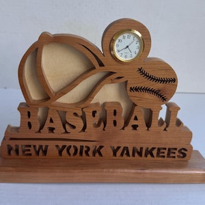 May include: Wooden baseball-themed desk clock featuring a baseball cap, baseball, and a small clock. The words "BASEBALL" and "NEW YORK YANKEES" are carved into the wood. The clock has a gold-colored frame and a white clock face.