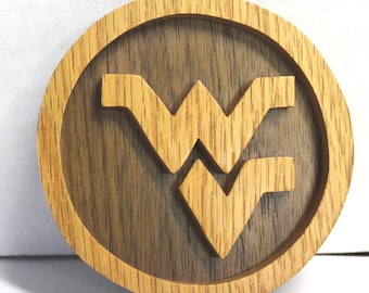 May include: A circular wooden ornament featuring a light-colored oak frame and a darker wood backing. The center displays a raised, light-colored wooden West Virginia University Mountaineers logo. A small hole is at the top for hanging.