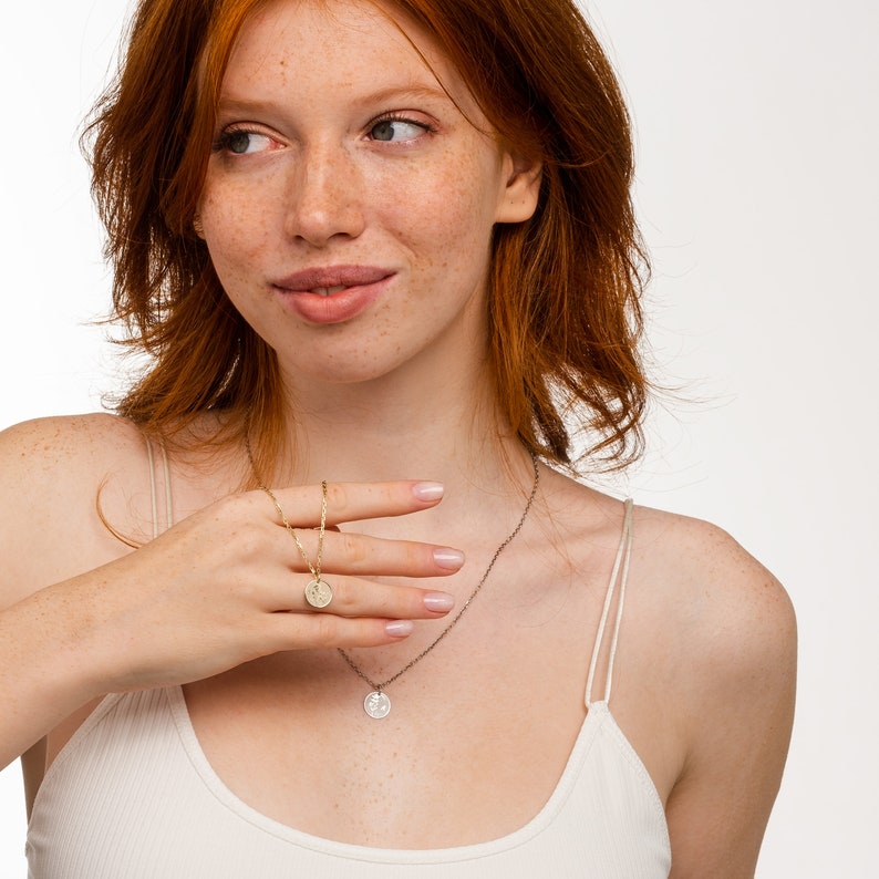 May include: A woman wearing a white tank top with two gold necklaces. One necklace has a small round pendant and the other has a larger round pendant.