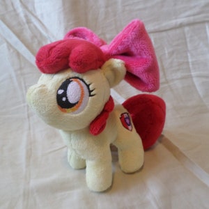 Può includere: Un giocattolo di peluche di Applejack di My Little Pony: L'amicizia è magia. Il giocattolo è color crema con un fiocco rosa e un simbolo di mela rossa sul suo fianco. Ha una criniera e una coda rosse.