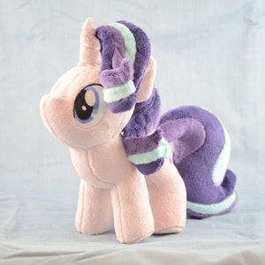 Può includere: Un giocattolo di peluche a forma di unicorno rosa con criniera e coda viola e blu chiaro. L'unicorno ha un occhio blu e un corno rosa.