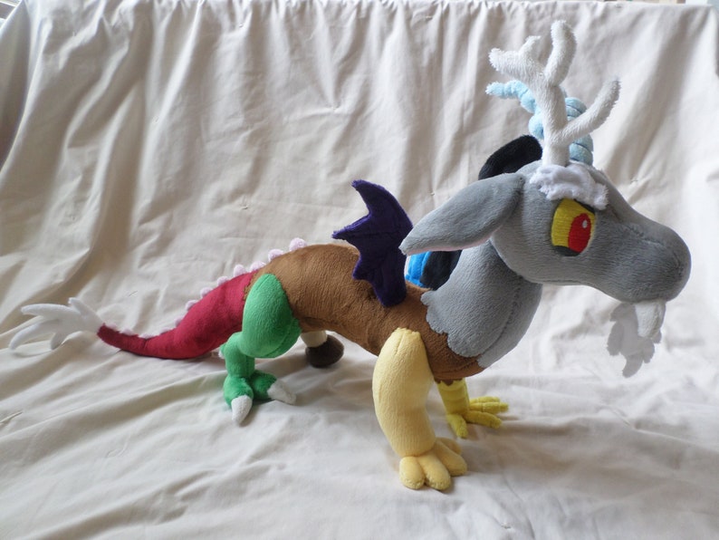 Pu&ograve; includere: Un giocattolo di peluche di Discord, un personaggio della serie animata My Little Pony: L'amicizia &egrave; magia. Discord &egrave; un drago grigio con un'ala viola, un corpo marrone, un braccio giallo, una gamba verde e una coda rossa. Ha corna bianche e una barba bianca.