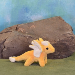 Può includere: Un piccolo drago di peluche giallo con ali bianche e una roccia marrone chiaro sullo sfondo.