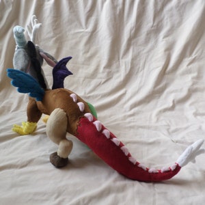 Pu&ograve; includere: Un drago di peluche con corpo marrone, coda rossa, ali blu e testa grigia con un'ala viola. Il drago ha accenti rosa sulla coda e sul corpo.