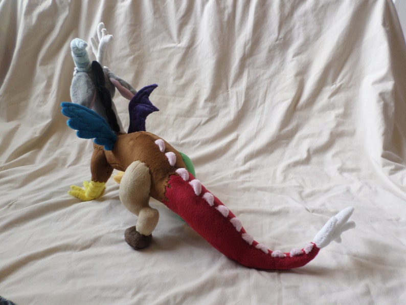 Pu&ograve; includere: Un drago di peluche con corpo marrone, coda rossa, ali blu e testa grigia con un'ala viola. Il drago ha accenti rosa sulla coda e sul corpo.